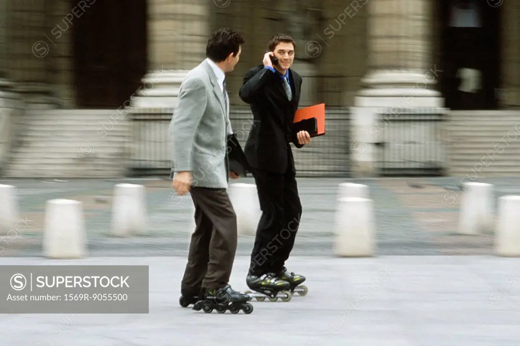 Men in business attire inline skating together along sidewalk, one phoning