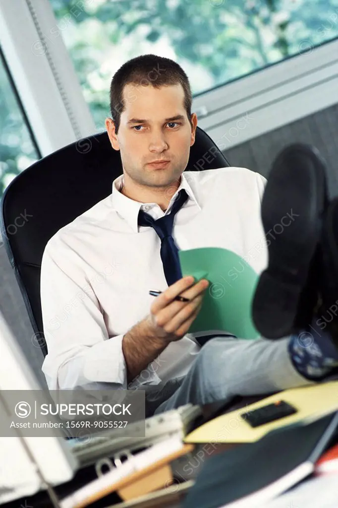Man with feet up on office desk holding document, looking away