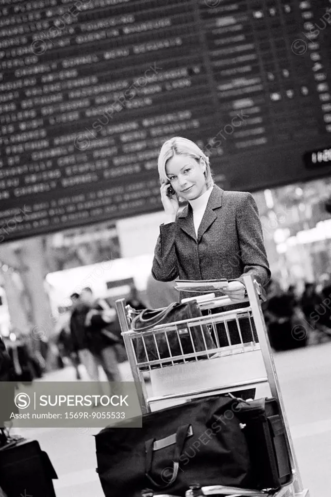 Business traveler phoning standing by luggage cart, arrival and departure board in background
