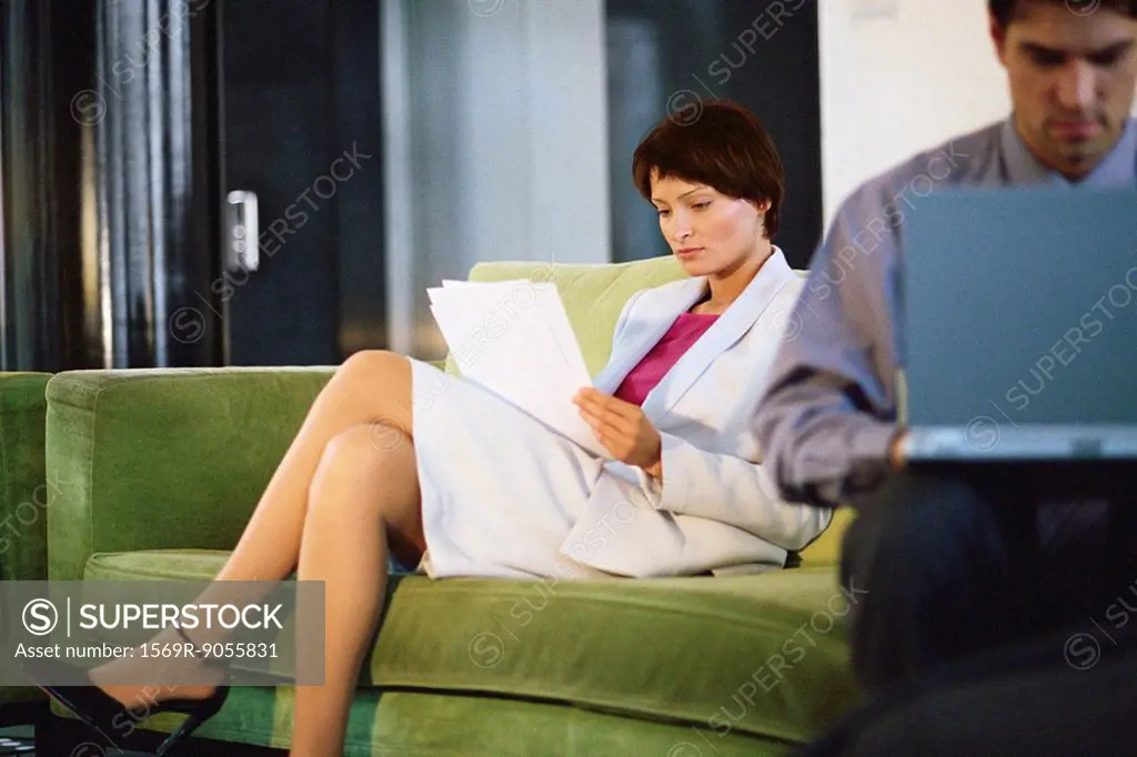 Female executive reading documents on sofa, man working on laptop in foreground
