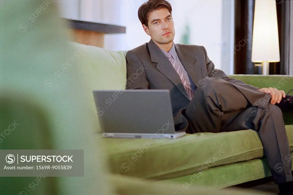 Businessman sitting on sofa with laptop, looking away