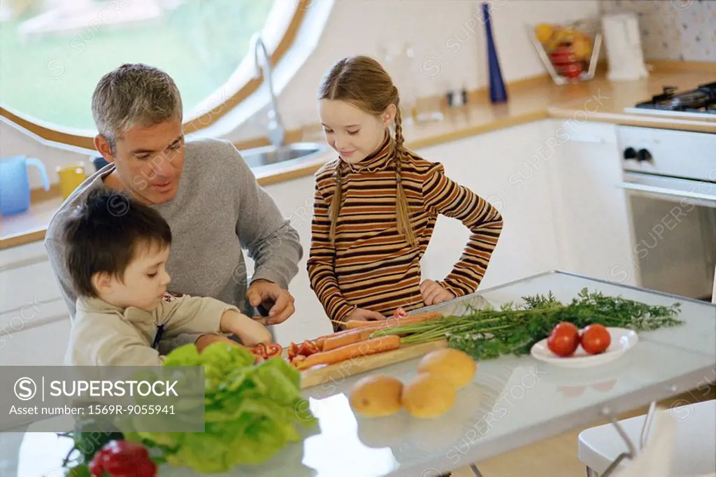 Father with children in kitchen, helping young son cut vegetables