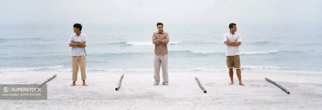 Three men with arms defiantly folded standing on beach, separated one from the other by lengths of wood