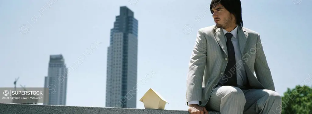 Man sitting on ledge beside miniature wooden house, skyscrapers in distance