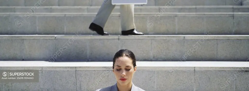 Woman on steps with eyes shut, businessman walking in background