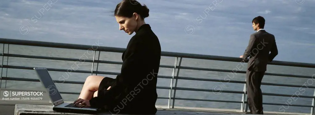 Businesswomen on waterfront bench using laptop computer, man at nearby railing looking at view