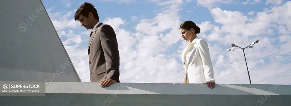 Businessman and businesswoman on rooftop, both looking down contemplatively