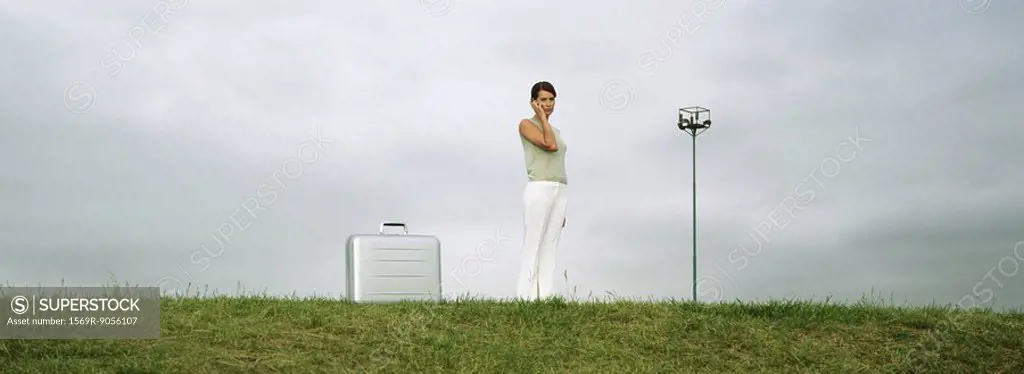 Woman standing in field using cell phone, briefcase on ground nearby