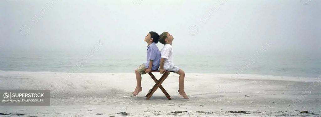 Two boys sitting back to back on chair on beach