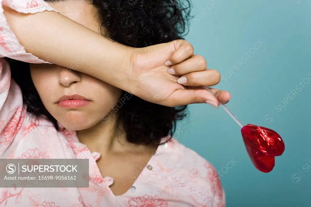 Woman frowning and covering eyes with arm, holding heart_shaped lollipop