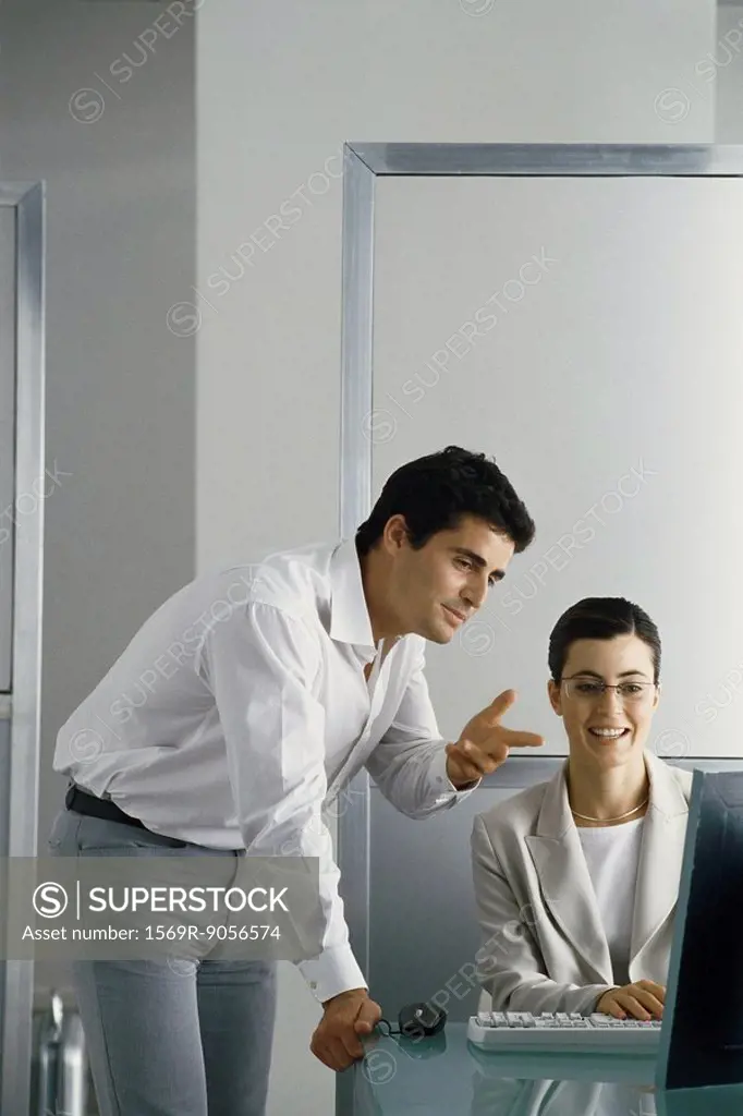Office scene, man leaning on edge of desk speaking to female colleague, both looking at her computer screen