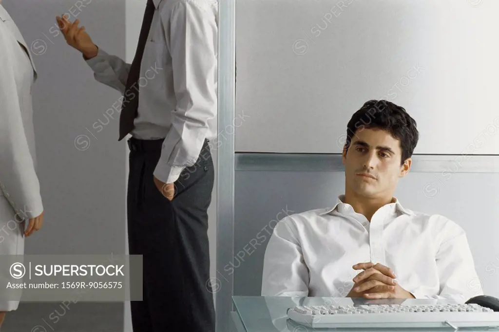 Man sitting at desk with hands clasped while colleagues talk nearby