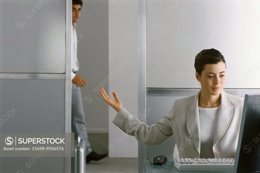 Businesswoman sitting at desk working, motioning for man to enter office