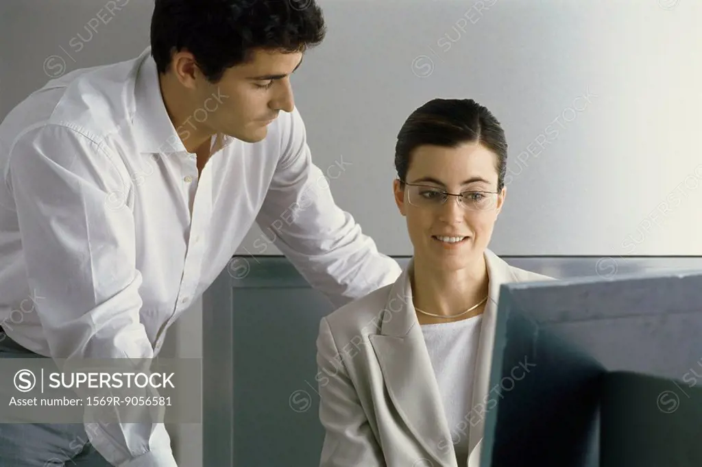 Young professional woman smiling, looking at computer screen while male colleague leans over her shoulder