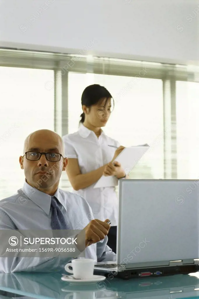 Businessman sitting at desk, looking at camera, female assistant taking notes in background