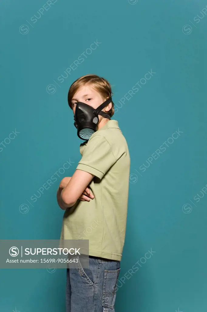 Boy wearing gas mask, arms folded, looking over shoulder at camera
