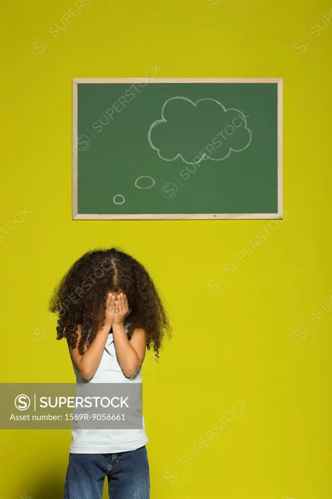 Little girl covering face with hands, thought bubble on chalkboard behind her