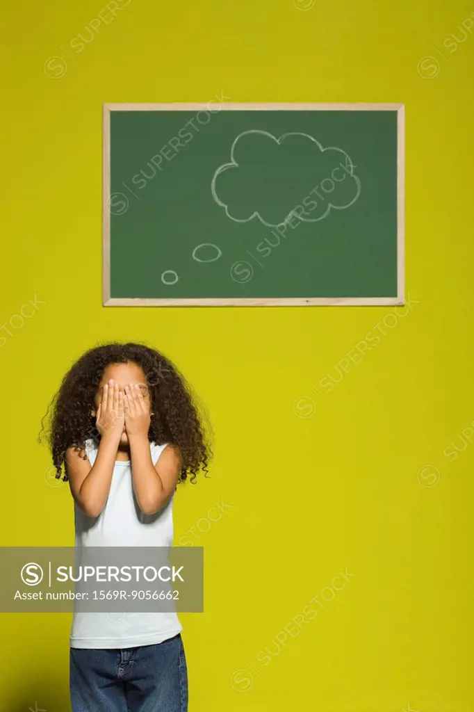 Little girl covering eyes with hands, thought bubble on chalkboard behind her