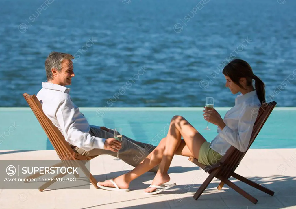 Couple sitting near sea in deck chairs, drinking champagne
