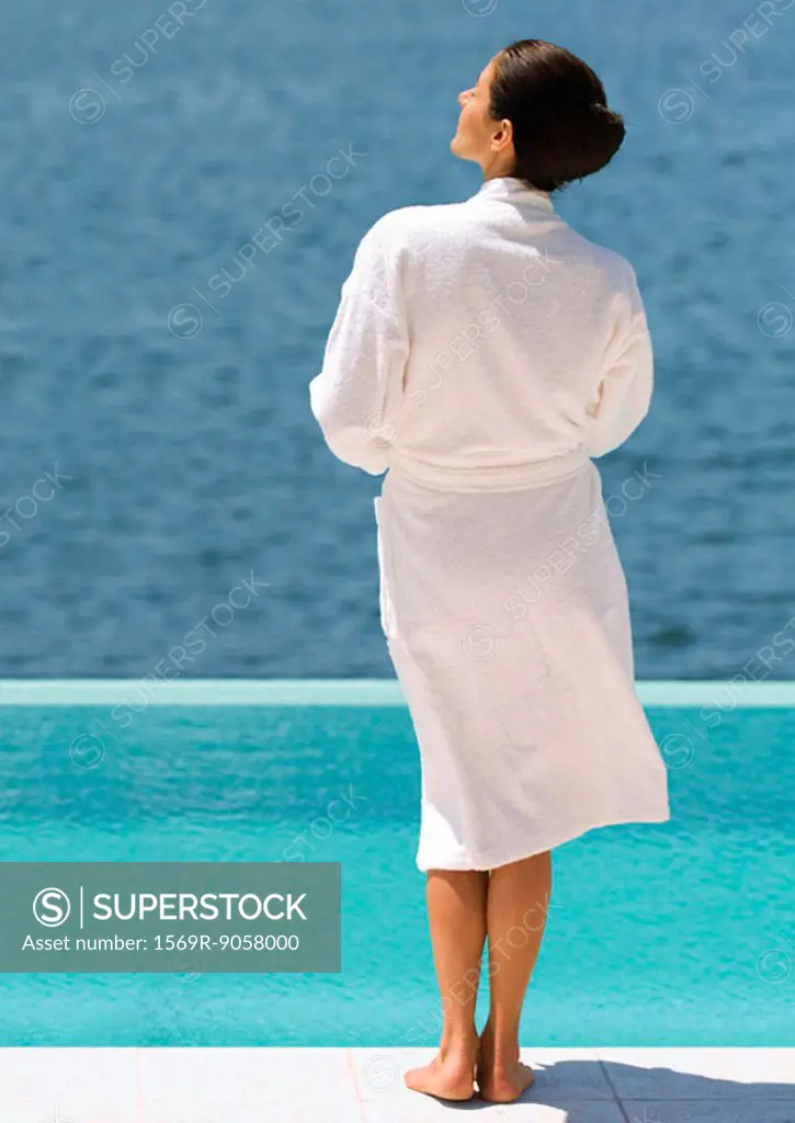 Woman in bathrobe standing near pool overlooking sea, rear view