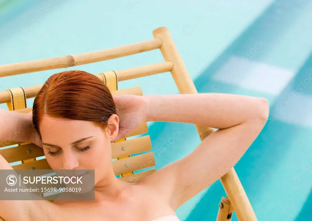 Woman sitting in deckchair with hands behind head and eyes closed, pool in background