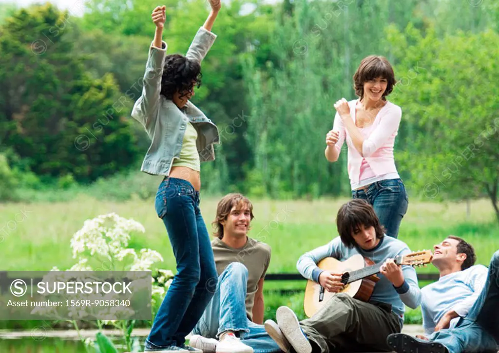 Group of friends outdoors, young women dancing while young man plays guitar