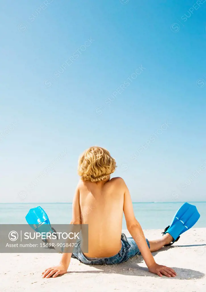 Boy wearing flippers sitting on beach, rear view