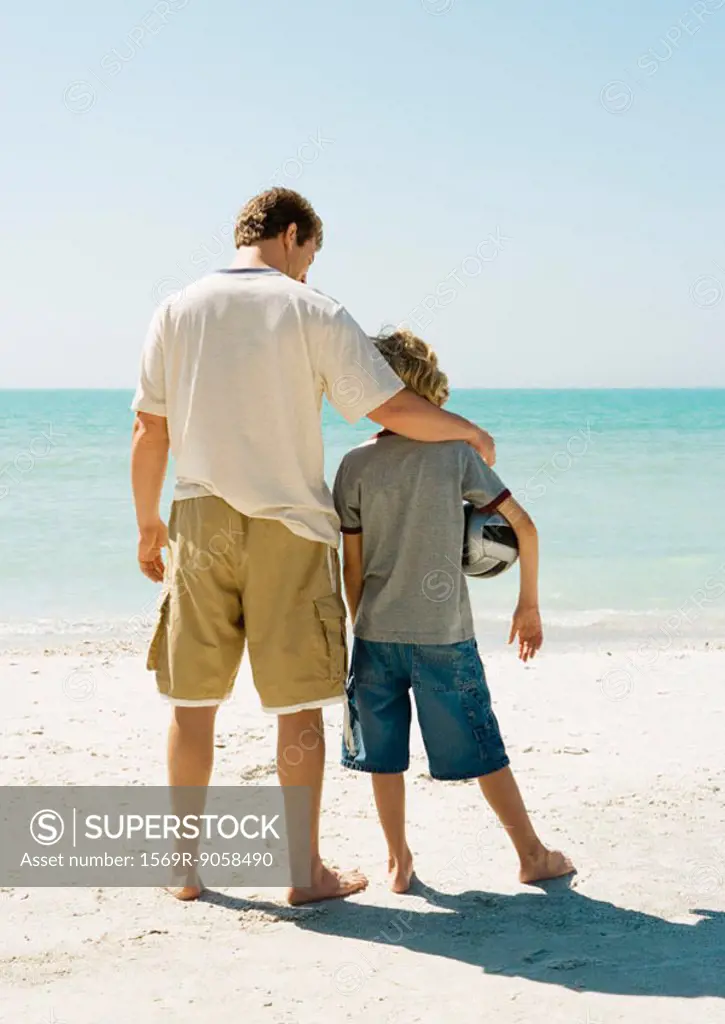 Father and son standing together on beach, boy holding ball under arm
