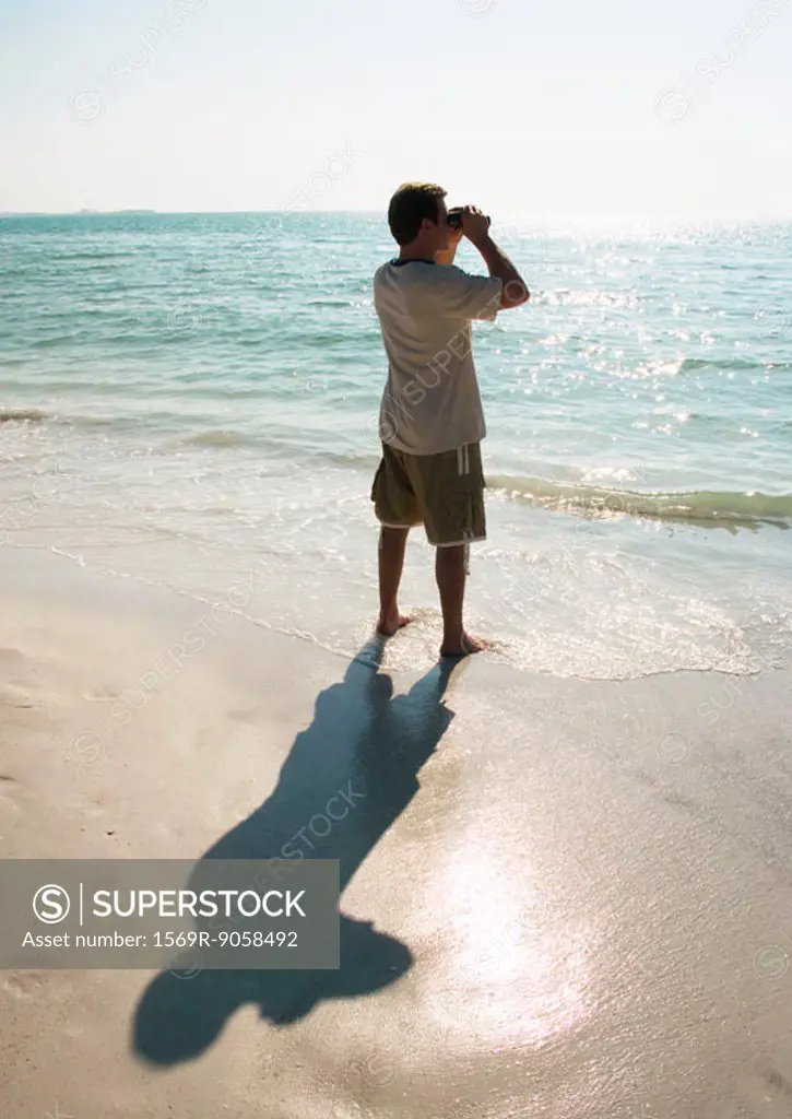 Man standing on beach, looking out to sea with binoculars