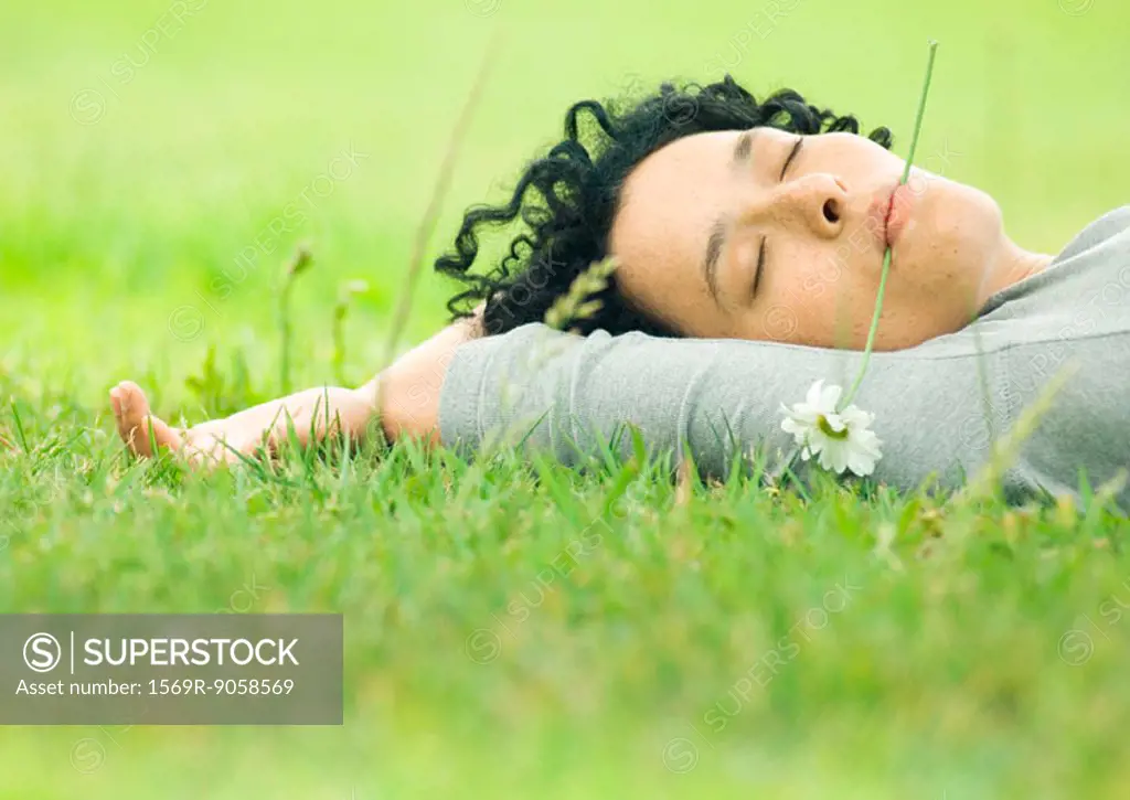 Young woman lying on grass with flower in mouth and eyes closed