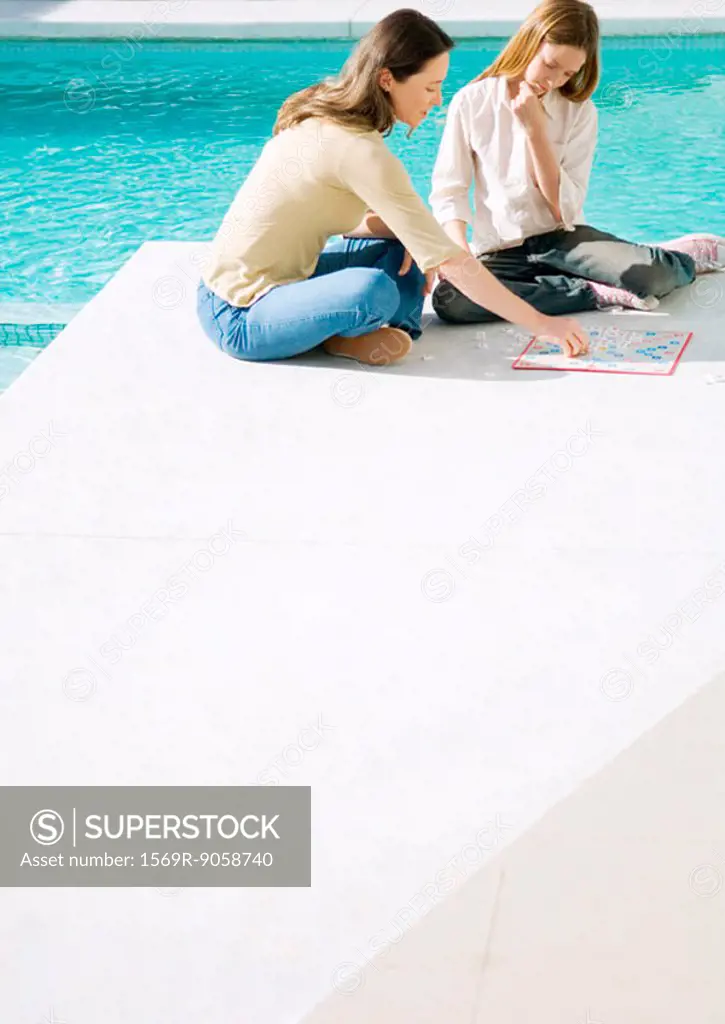Mother and daughter sitting by edge of pool, playing board game