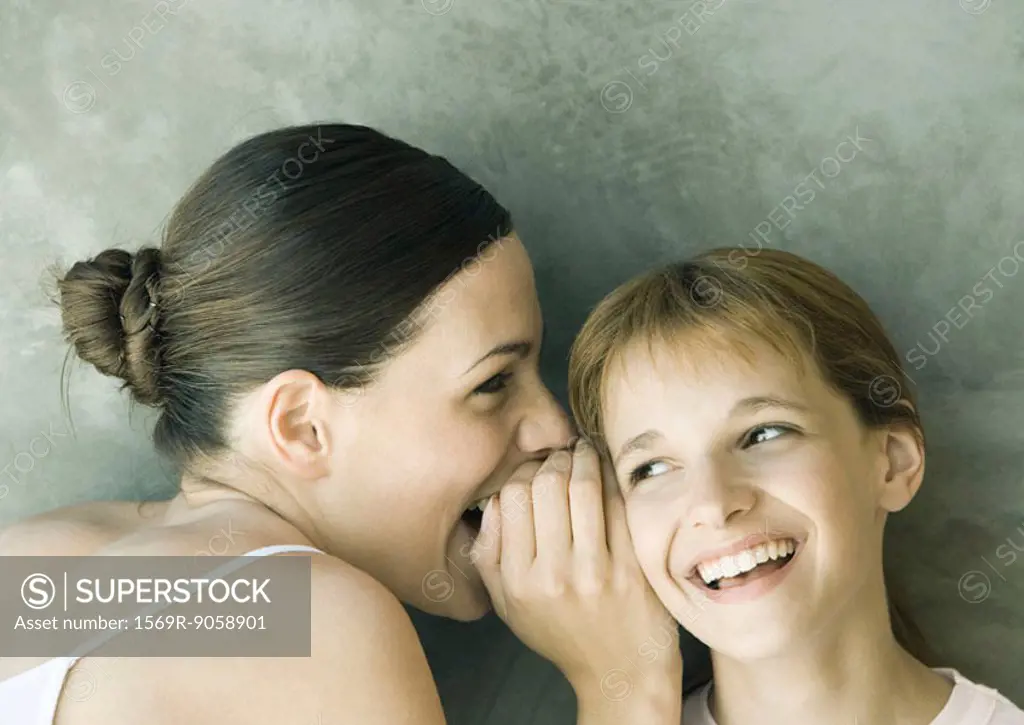 Young woman whispering to girl, both laughing, portrait, close-up
