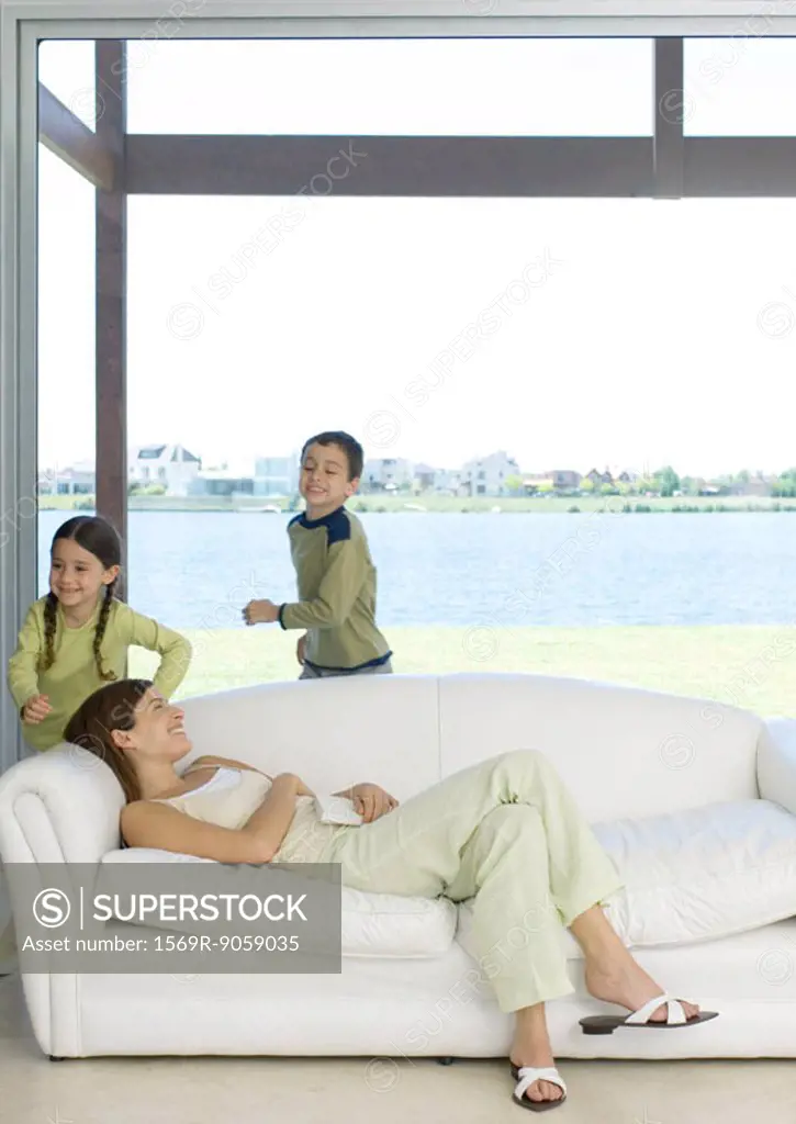 Mother reclining on sofa while children run around her