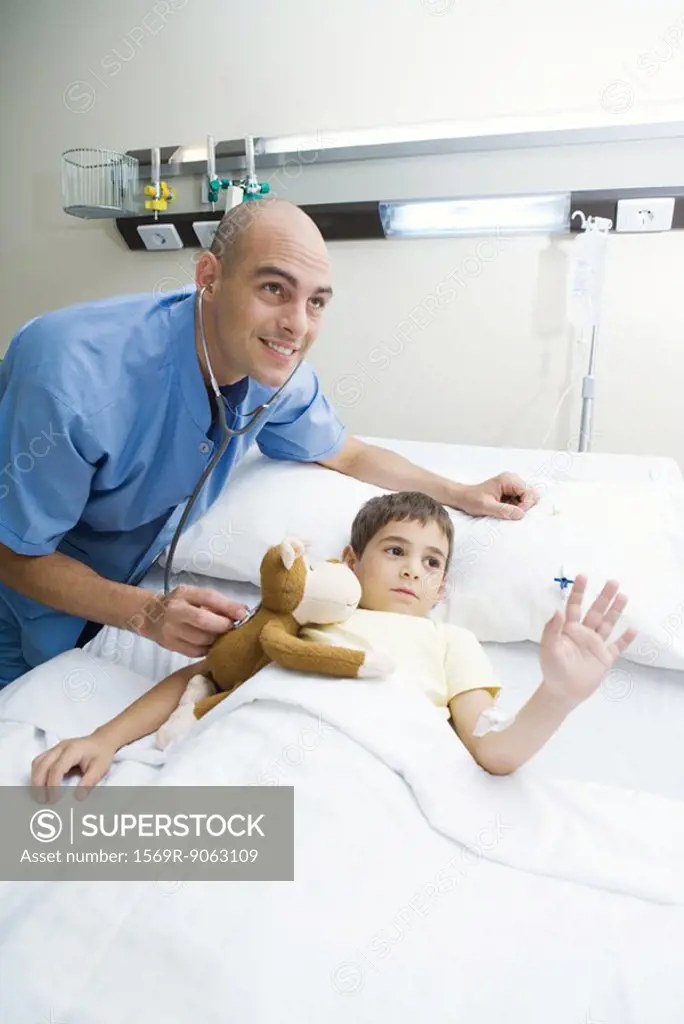 Boy lying in hospital bed, intern holding stethoscope to boy´s stuffed animal, both looking out of frame