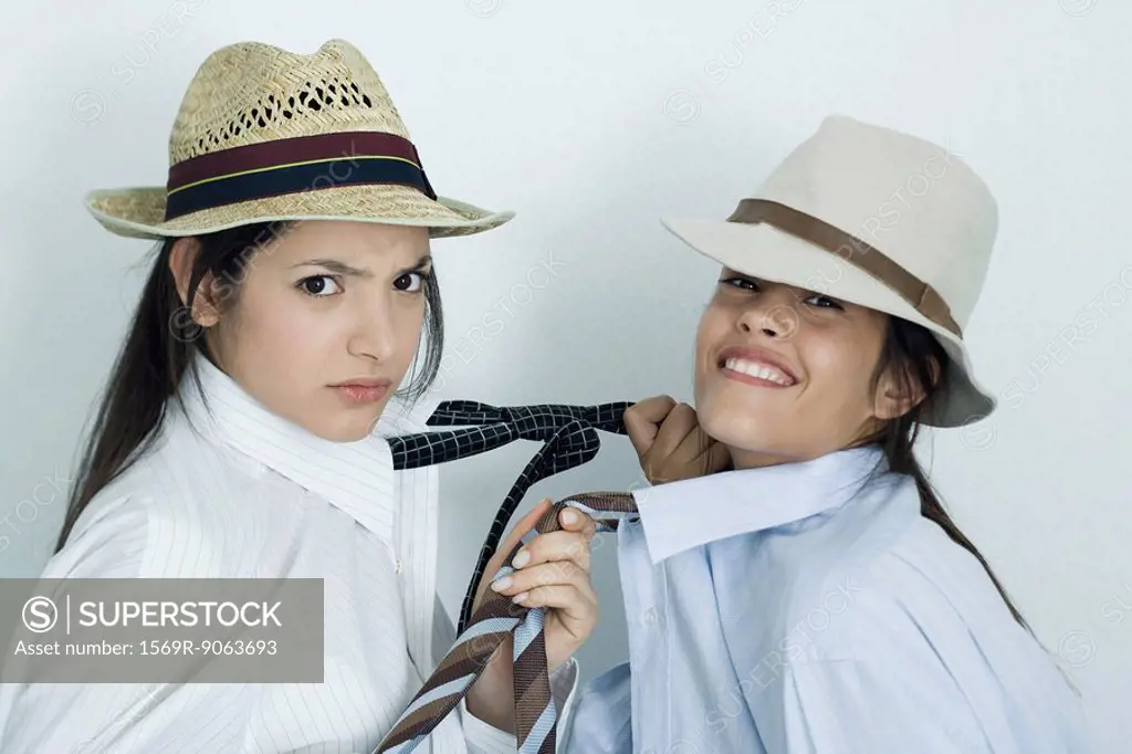 Two young female friends dressed in button down shirts, ties and hats, pulling on each other´s ties, portrait