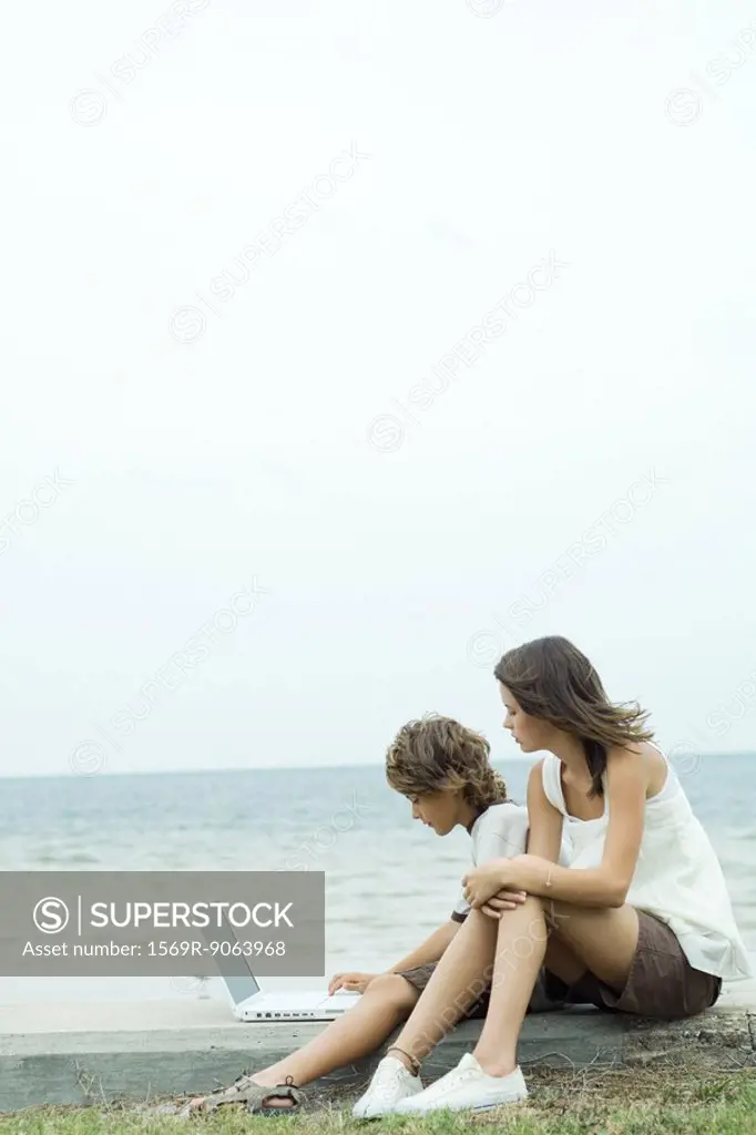 Boy and teen sister using laptop by water´s edge