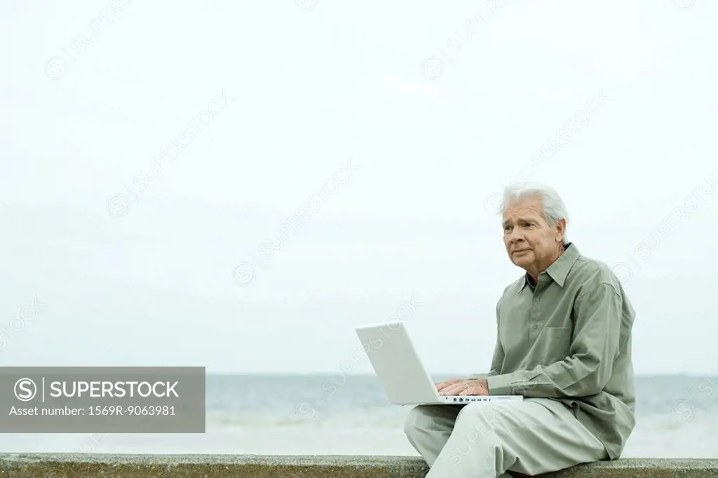 Senior man sitting near water´s edge, using laptop