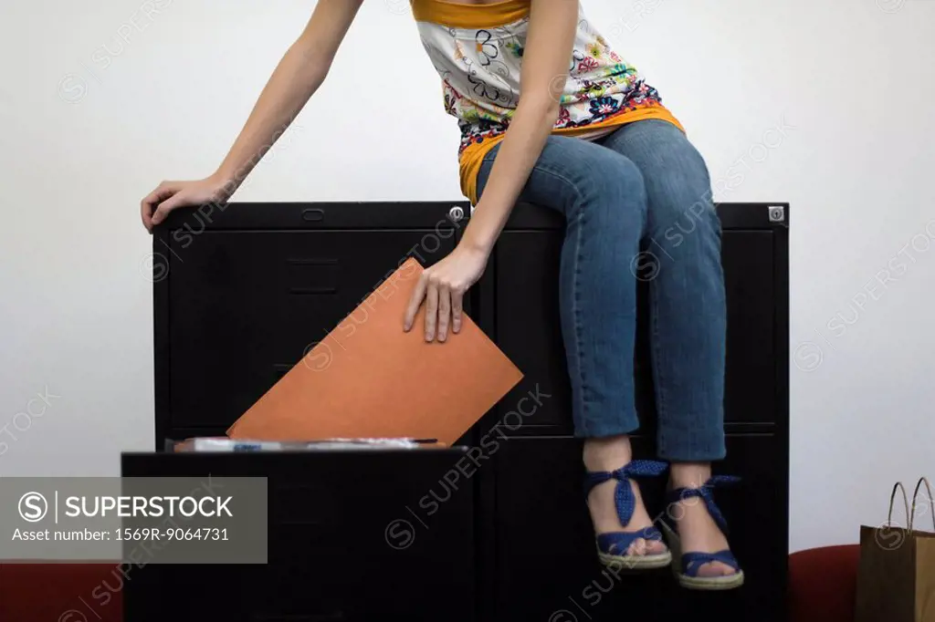 Woman sitting on filling cabinets, removing file from drawer, cropped