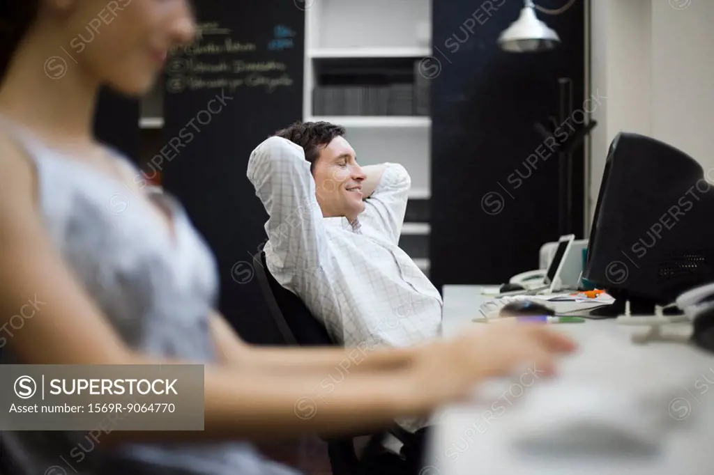Man relaxing at desk, woman working in foreground