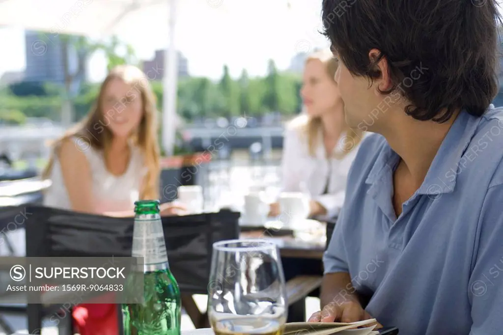 Young man sitting at outside cafe, looking over shoulder at young women sitting behind him