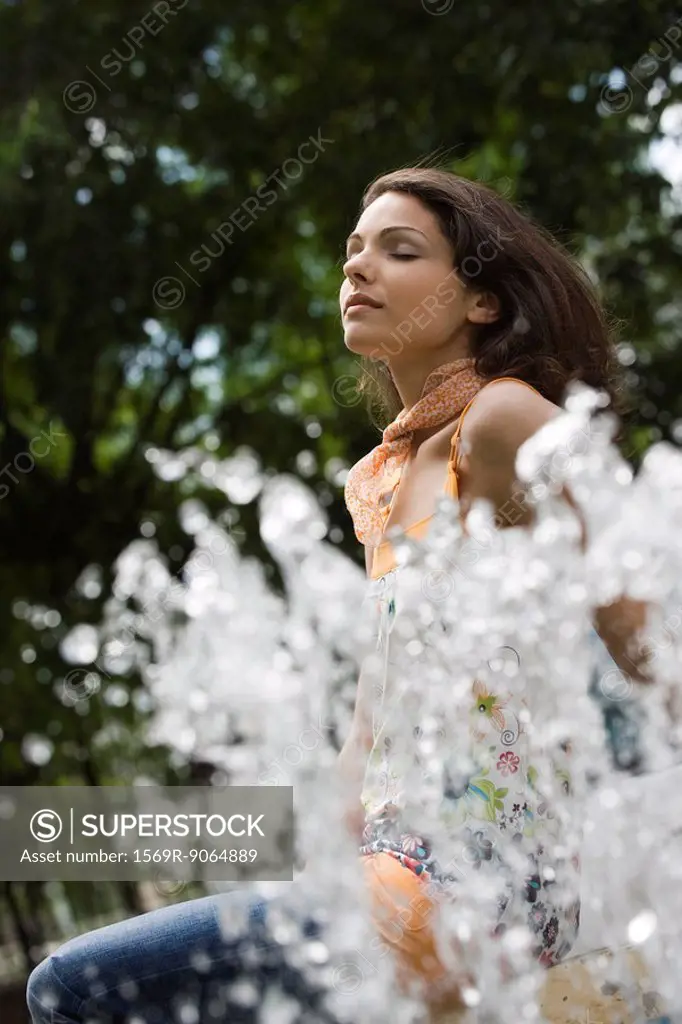 Young woman sitting by outdoor fountain with eyes closed, low angle view