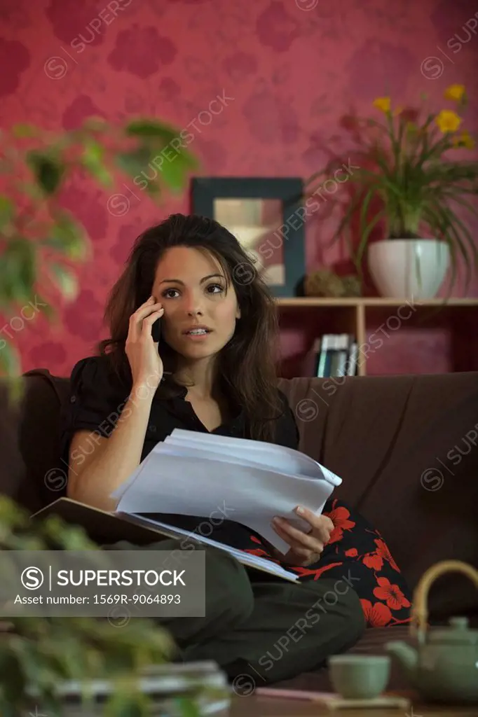 Woman sitting on sofa, using cell phone and holding document