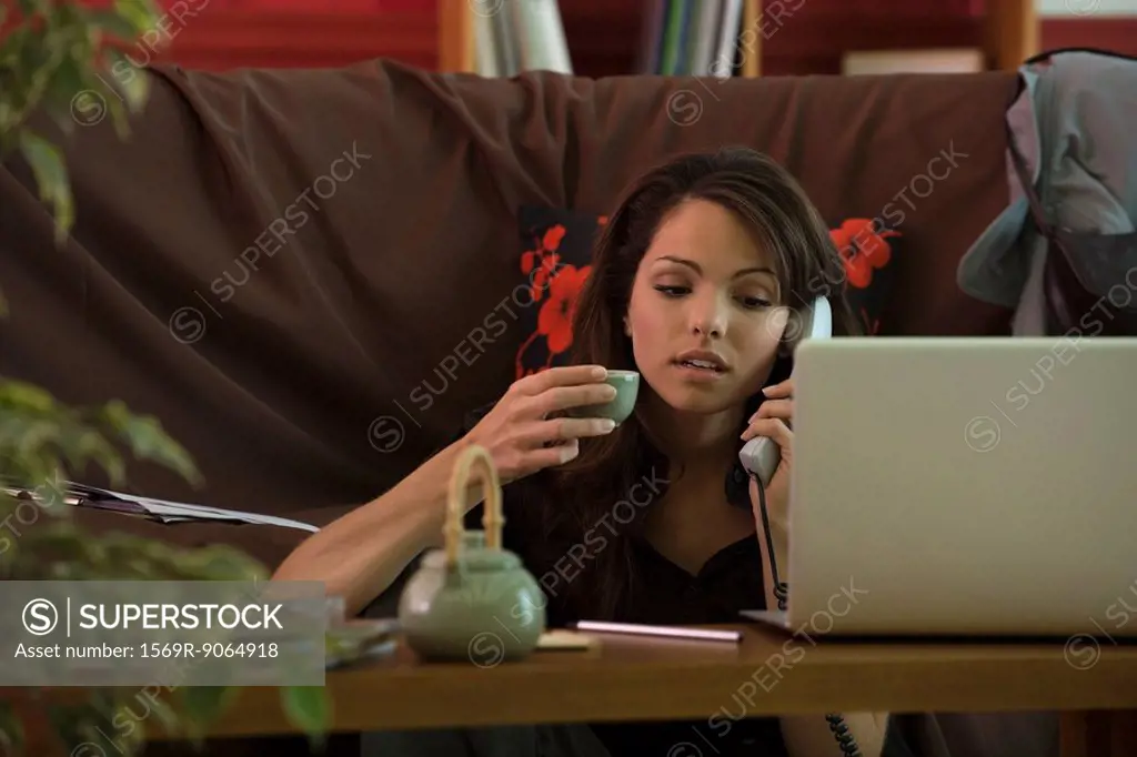 Young woman sitting at coffee table using phone and laptop computer, holding cup of tea