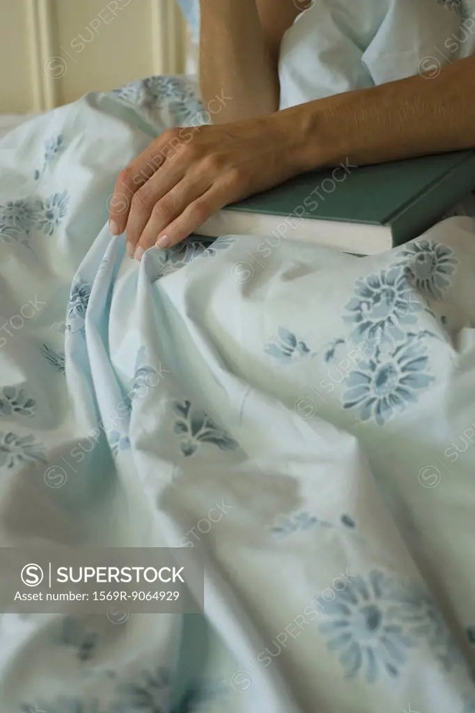 Woman sitting in bed with hand resting on book, cropped view