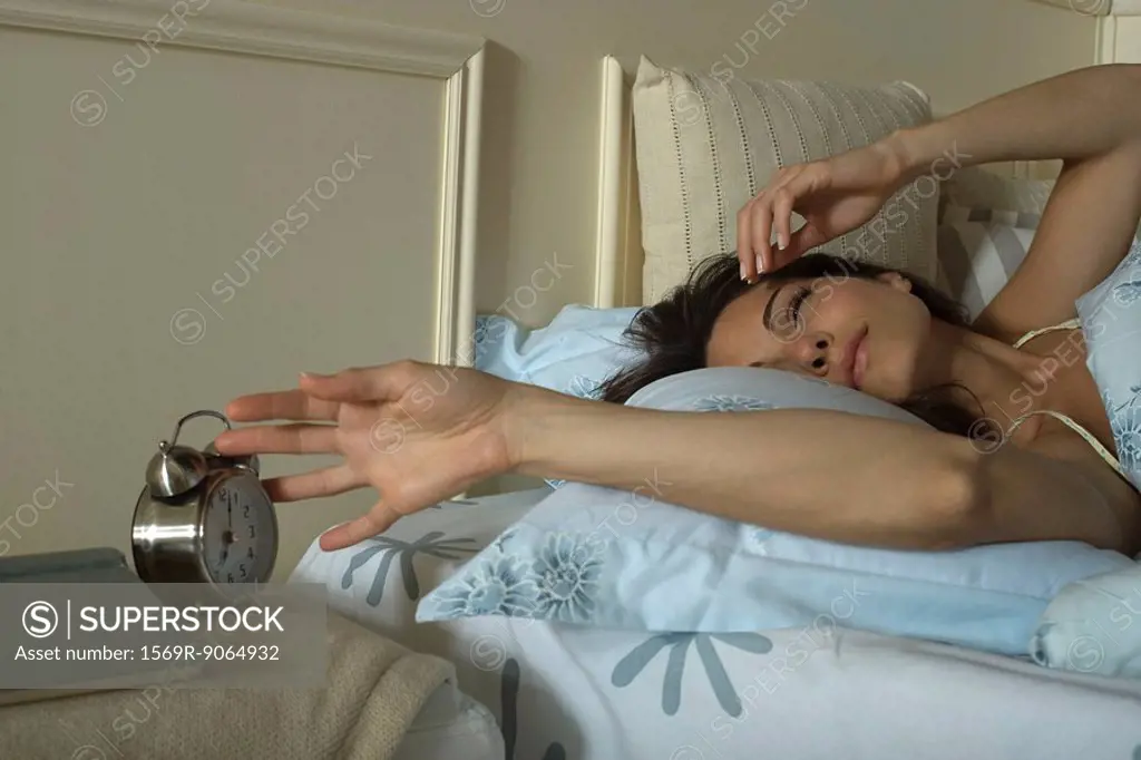 Young woman lying in bed, silencing alarm clock