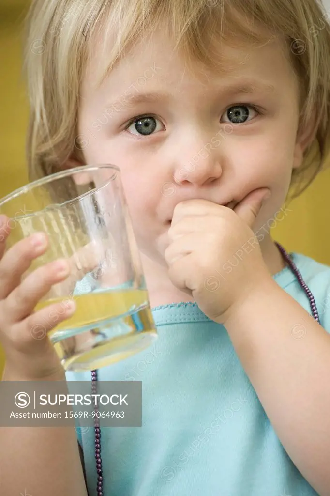Toddler girl holding glass of juice, covering mouth with one hand