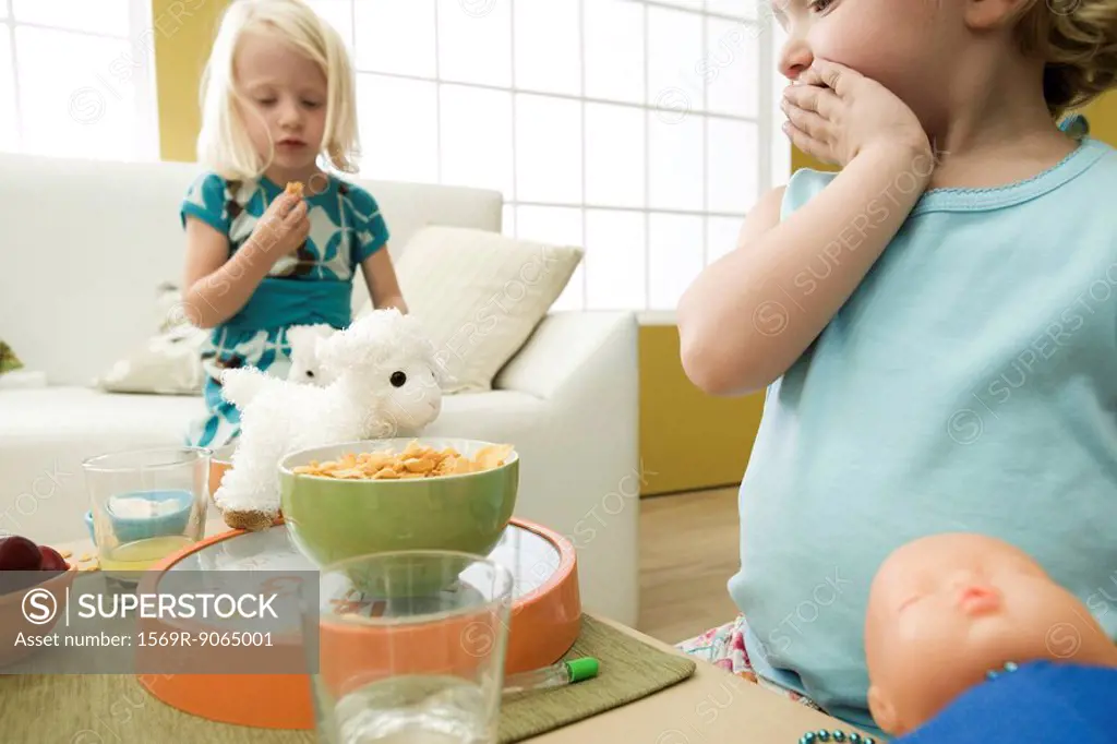 Little girl looking at bowl of cereal with hand over mouth, sister in background