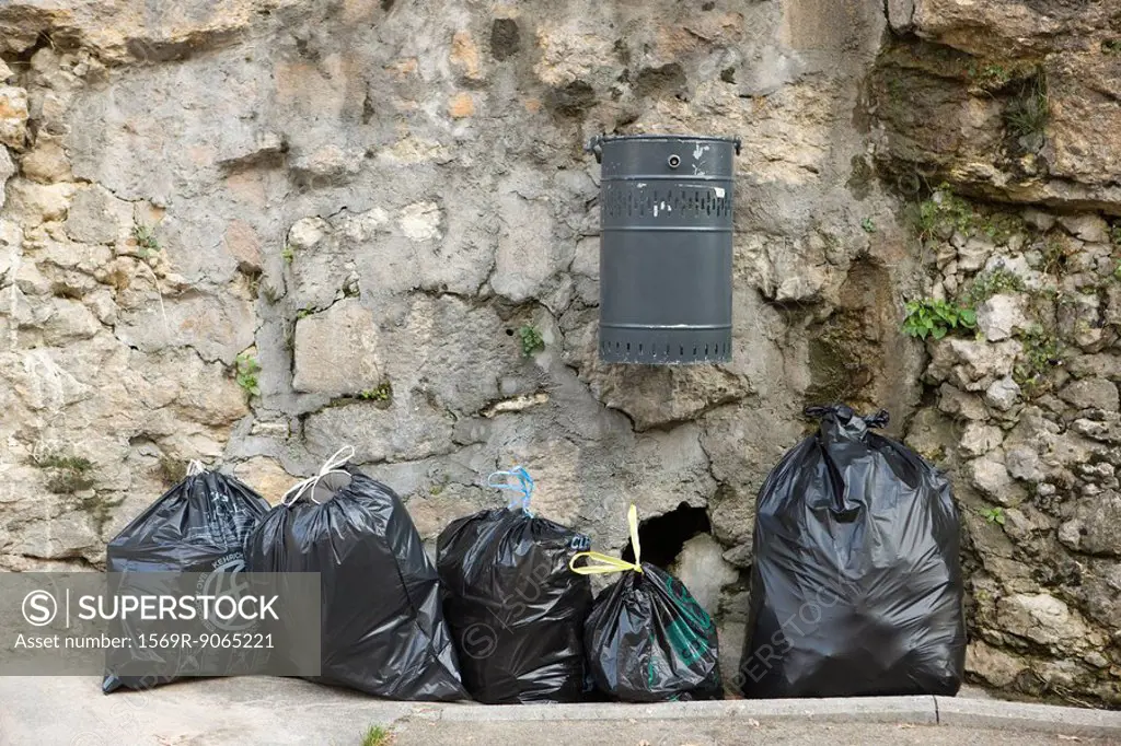 Several bags of garbage lined up along stone wall beneath mounted trash can