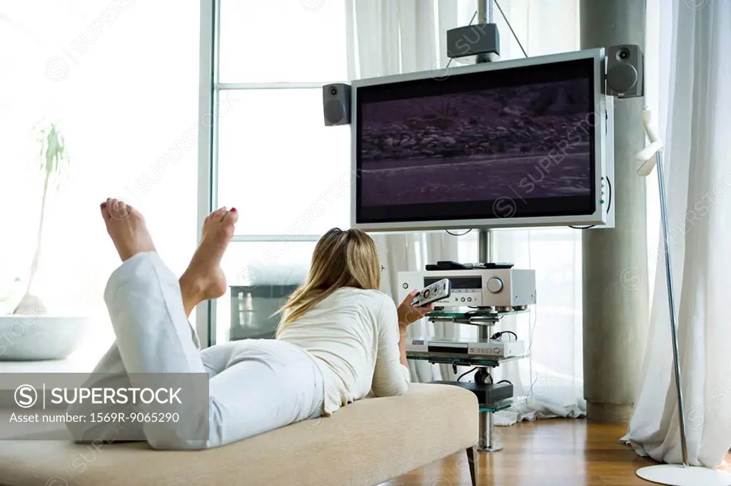 Female lying on chaise longue, watching flat screen television with surround sound