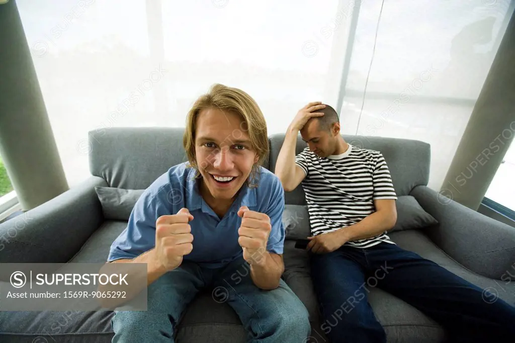 Two men sitting on sofa watching television, one cheering, other holding head